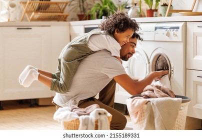 Side view of black child in casual clothes with curly hair smiling and embracing dad loading washing machine during household routine in morning at home - Shutterstock ID 2155189331