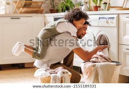Side view of black child in casual clothes with curly hair smiling and embracing dad loading washing machine during household routine in morning at home