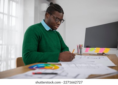 Side View Of Black Designer Man Drawing Sitting At Desk Covered With Papers Working Via Pencil And Ruler Creating Plan In Office  Modern Architecture And Design Career Concept