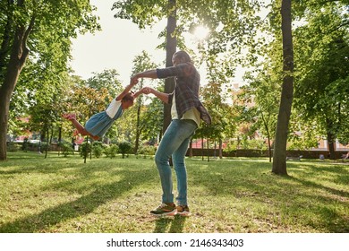 Side View Of Black Father Holding And Spinning High In Hands His Joyful Little Daughter On Lawn In Green Park. Family Relationship And Spending Time Together. Fatherhood And Parenting. Sunny Daytime