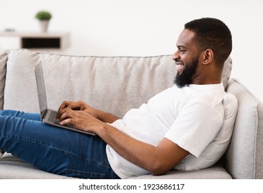 Side View Of Black Freelancer Guy Working On Laptop Computer Lying On Sofa And Browsing Internet Working Remotely From Home. People, Technology And Freelance Lifestyle Concept