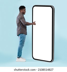Side View Of Black Guy Using Big Cellphone With Blank White Screen Touching Huge Display Panel With Finger, Cheerful Man Standing On Blue Background, Ordering Food Delivery, Mock Up. Full Body Length - Shutterstock ID 2108514827