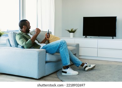 Side View Of Black Guy Watching Sports Game On TV With Blank Screen, Celebrating Victory And Shaking Fists, Sitting On Sofa In Living Room. Mockup