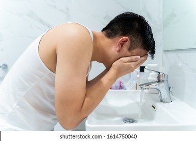 Side View Of Black Haired Asian Man In White Shirt Leaning Over Sink While Washing Face In Bathroom