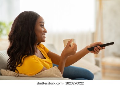 Side View Of Black Lady With Remote Control And Coffee Watching TV At Home. Millennial African American Woman With Television Controller Drinking Aromatic Beverage And Switching Channels In Morning