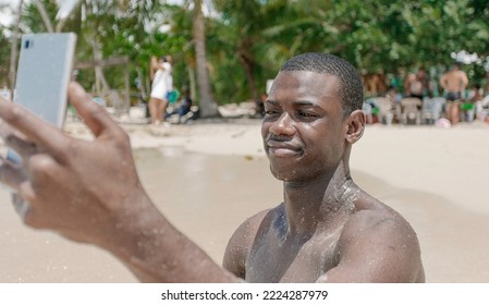 Side View Black Male Traveler Sitting On The Sand Near The Sea And Taking A Selfie During Summer Vacation At The Resort.