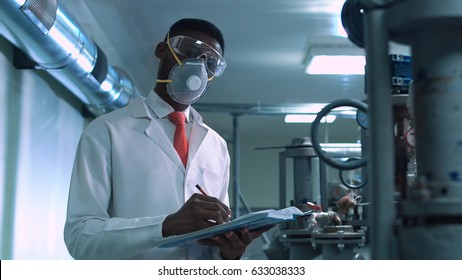 Side View Of Black Scientist In White Lab Coat, Goggles And Mask Standing Next To Productional Equipment With Journal And Writing
