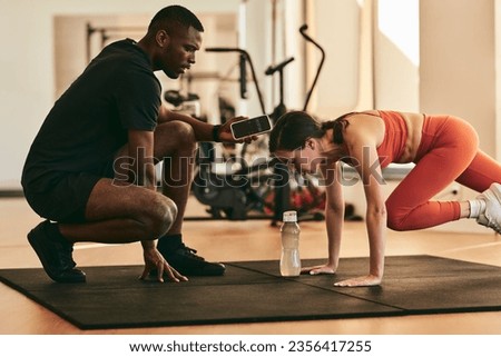 Side view of black trainer using smartphone with timer while sportswoman doing mountain climber exercise during High Intensity Interval Training at gym
