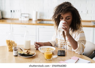 The side view of a black woman drinking from a cup and having breakfast at the table in the bright kitchen