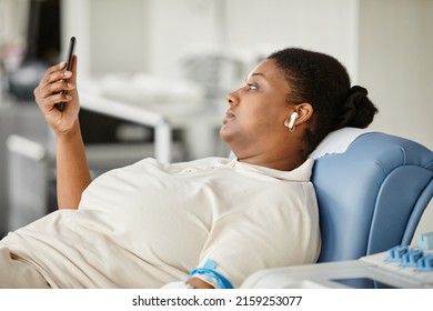 Side View Of Black Young Woman Giving Blood While Laying In Chair At Blood Donation Center And Holding Smartphone