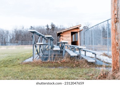 Side view of bleacher seats and a baseball dugout with trees in the background and cloudy skies - Shutterstock ID 2312047659