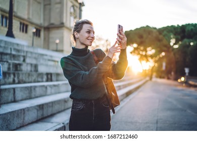 Side View Of Blonde Female Taking Selfie With Mobile Phone Near Steps Of Aged Town Building Against Bright Back Lit 