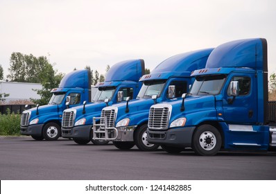 Side view of the blue big rigs semi trucks tractors with high roof spoiler for better aerodynamic flow stand in row without semi trailers on the parking lot and waiting for loading cargo for delivery