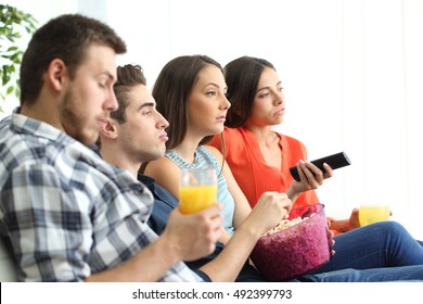 Side View Of Bored Group Of Four Friends Watching A Bad Tv Program Sitting On A Sofa In The Livingroom Of An Apartment