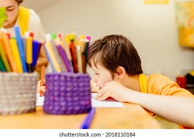 Side View Of Boy Child Down Syndrome In Orange Shirt Drawing With Pencil On Large White Sheet Of Paper In The Classroom With Different Pencils In Crate. Art Therapy For Children Special Needs Concept.