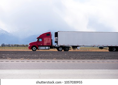 Side view of bright red big rig semi truck fleet transporting cargo in long refer semi trailer on the flat road in Utah with snow covered mountain and clouds on background