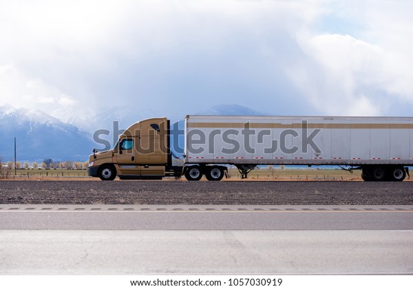 Side view of brown big\
rig semi truck fleet transporting cargo in long dry van semi\
trailer on the flat road in Utah with snow covered mountain and\
clouds on background