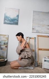 Side view of brunette artist in sweater and bra holding paintbrushes near drawings in workshop - Shutterstock ID 2251005245