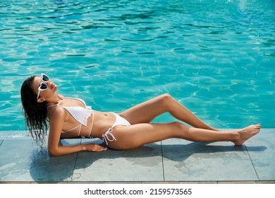 Side view of brunette girl with long wet hair lying near swimming pool. Slim young female in sunglasses sunbathing, relaxing, looking up. Concept of summertime and youth.