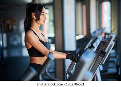 Side View Of Brunette Woman In Black Sportswear And Wireless Headphones Doing Hard Cardio Workout On Orbitrek. Sexy Muscular Female In Gloves, With Slim Body Running In Empty Gym. Concept Of Sport.