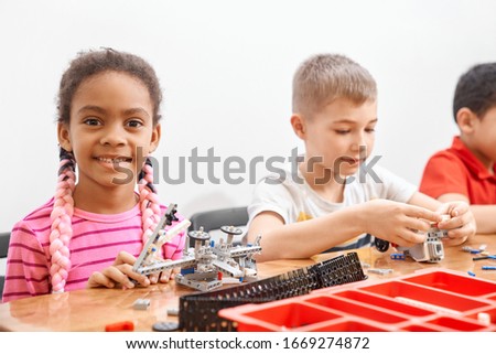 Side view of building kit for group of three multiracial kids creating toys, having positive emotions and joy, african girl smiling and looking at camera. Close up of friends working on project.