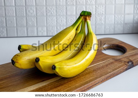 Side view of a bunch of yellow bananas which are laying on a wooden cutting board against background of white ceramic tiles