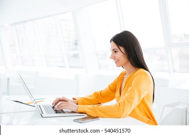 Side View Of Business Woman In Orange Shirt Using Laptop Computer And Sitting By The Table In Office