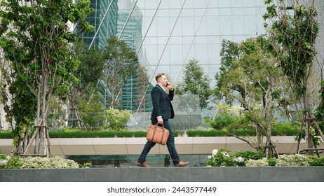 Side view of businessman calling manager about marketing report and walking at modern building in green eco city. Leader going to workplace while talking to his team by using phone. Lifestyle. Urbane. - Powered by Shutterstock