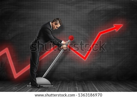 Side view of businessman looking down and pulling big lever near black graph ruled wall with red graph arrow on it. Jumpstart your business. Work hard. Achieve success.