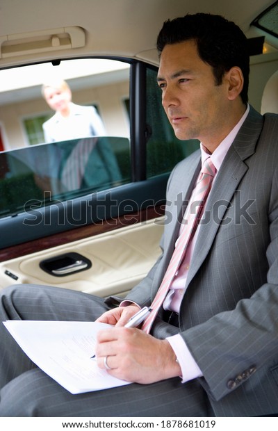 Side view of a businessman sitting in the backseat\
of the car and thinking while holding a pen and document on the\
lap.