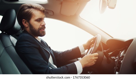 Side View Of Businessman Sitting In Car Behind Wheel Looking At Smartphone On Dashboard And Talking