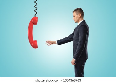 Side View Of Businessman Standing And Reaching Out For Big Red Landline Phone Receiver Dangling Down On Wire From Above. Get Useful Advice. Help People In Trouble. Consulting Business.