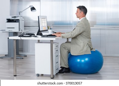 Side View Of Businessman Using Computer While Sitting On Exercise Ball At Office