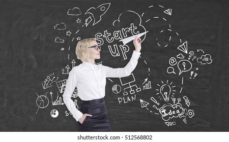 Side view of businesswoman with short blond hair wearing glasses who is throwing paper plane near blackboard with project launch sketches