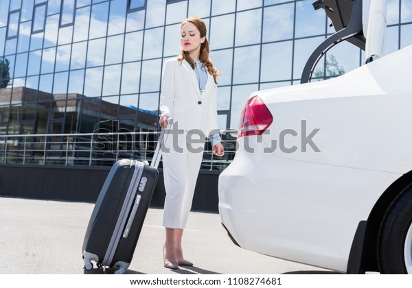 side view of businesswoman in white suit with\
suitcase standing at car on\
street