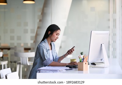 Side view of businesswoman working with computer and using smart phone while sitting in modern office. - Shutterstock ID 1892683129
