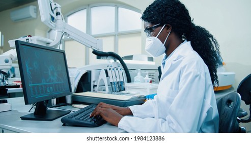 Side view of busy African American young female medical researcher sitting in lab working typing on computer looking at molecular structure developing covid cure. Modern laboratory, clinic research
