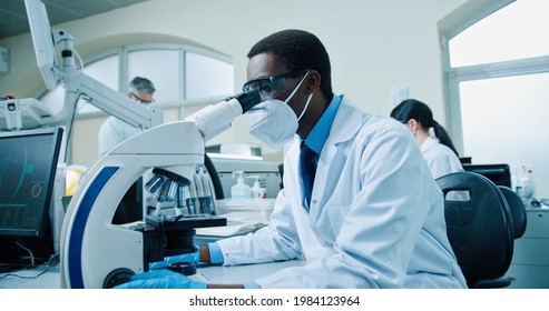 Side view of busy African American young professional male scientist medic in mask sitting at laboratory working on modern technology looking in microscope working on cure, experiment analysis concept