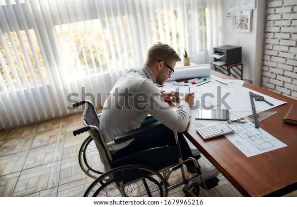 Side view of busy male engineer in a\
wheelchair sketching a construction project while working at his\
workplace in the modern office. Disability and handicap concept.\
Construction and\
architecture
