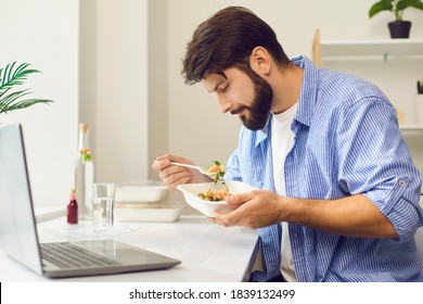 Side View Of Busy Young Man In Casual Wear Sitting At Desk At Home Or In The Office And Eating Takeaway Food From Containers During Lunch Break. Healthy Meal In Workplace Concept