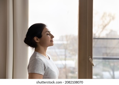 Side View Calm Happy Inspired Beautiful Young Indian Ethnicity Woman Standing Near Window, Breathing Fresh Air, Meditating With Closed Eyes, Imagining Good Future, Enjoying Carefree Leisure Time.