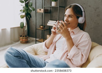 Side view calm young woman wears casual clothes sits in armchair listen to music in headphones drink coffee stay home hotel flat rest relax spend free spare time in living room indoor. Lounge concept - Powered by Shutterstock