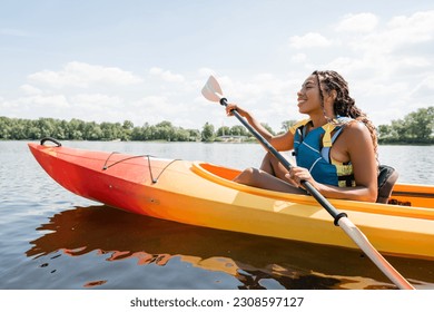 side view of carefree and active african american woman in life vest holding paddle while sailing in kayak on lake with green picturesque shore in summer - Powered by Shutterstock