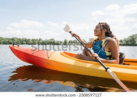 side view of carefree and active african american woman in life vest holding paddle while sailing in kayak on lake with green picturesque shore in summer