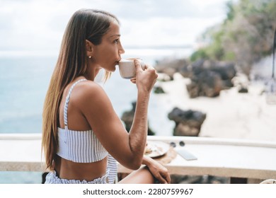 Side view of carefree female freelancer sitting at table and drinking coffee while admiring nature in cafe at resort during summer holidays in Bali - Powered by Shutterstock