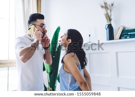 Side view of carefree woman standing at home and putting put tongue while man having important conversation on smartphone and keeping finger on lips