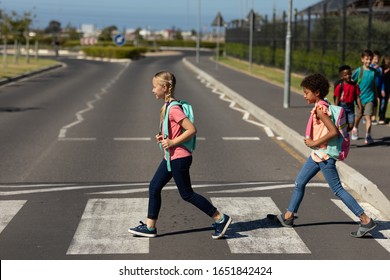 Side view of a Caucasian and an African American schoolgirl wearing rucksacks crossing the road at a pedestrian crossing on their way to elementary school on a sunny day, with other schoolchildren in - Powered by Shutterstock