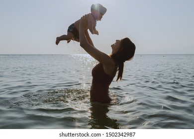 Side View Of Caucasian Attractive Mom Lifting Her 6 Months Old Son While Standing In Water. Baby Having Cap On Head And Looking At Mother.