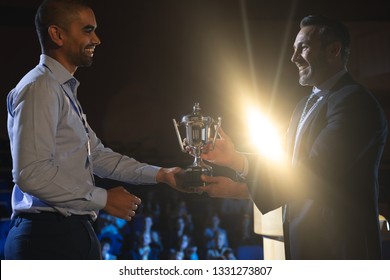 Side view of Caucasian businessman giving trophy to mixed race business male executive on stage in auditorium