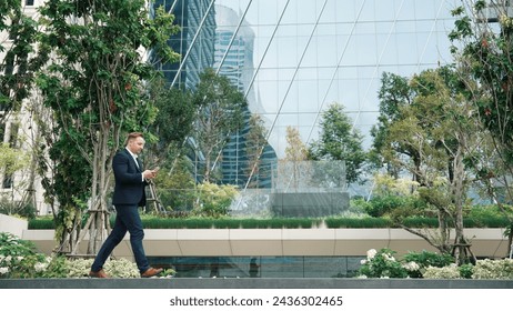 Side view of caucasian businessman looking at smart phone and walking at modern building in green eco city. Skilled leader going to workplace while checking report by using phone. Lifestyle. Urbane. - Powered by Shutterstock